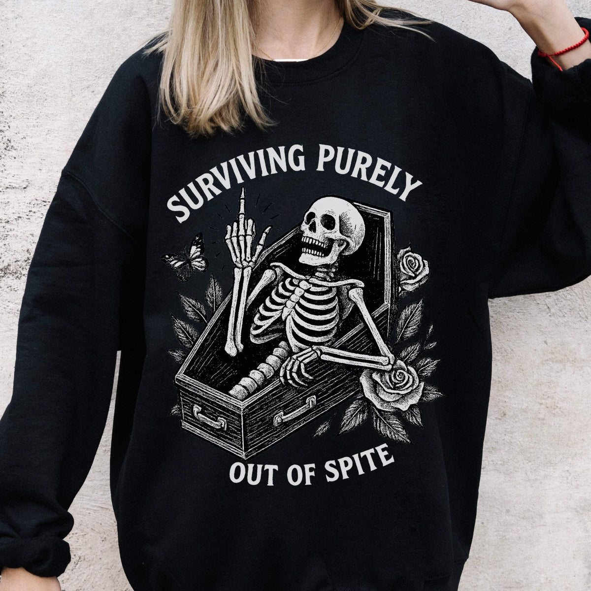 May include: Black sweatshirt with a white graphic of a skeleton in a coffin, giving the middle finger. The text reads "SURVIVING PURELY OUT OF SPITE". Roses and a butterfly are also featured in the design.
