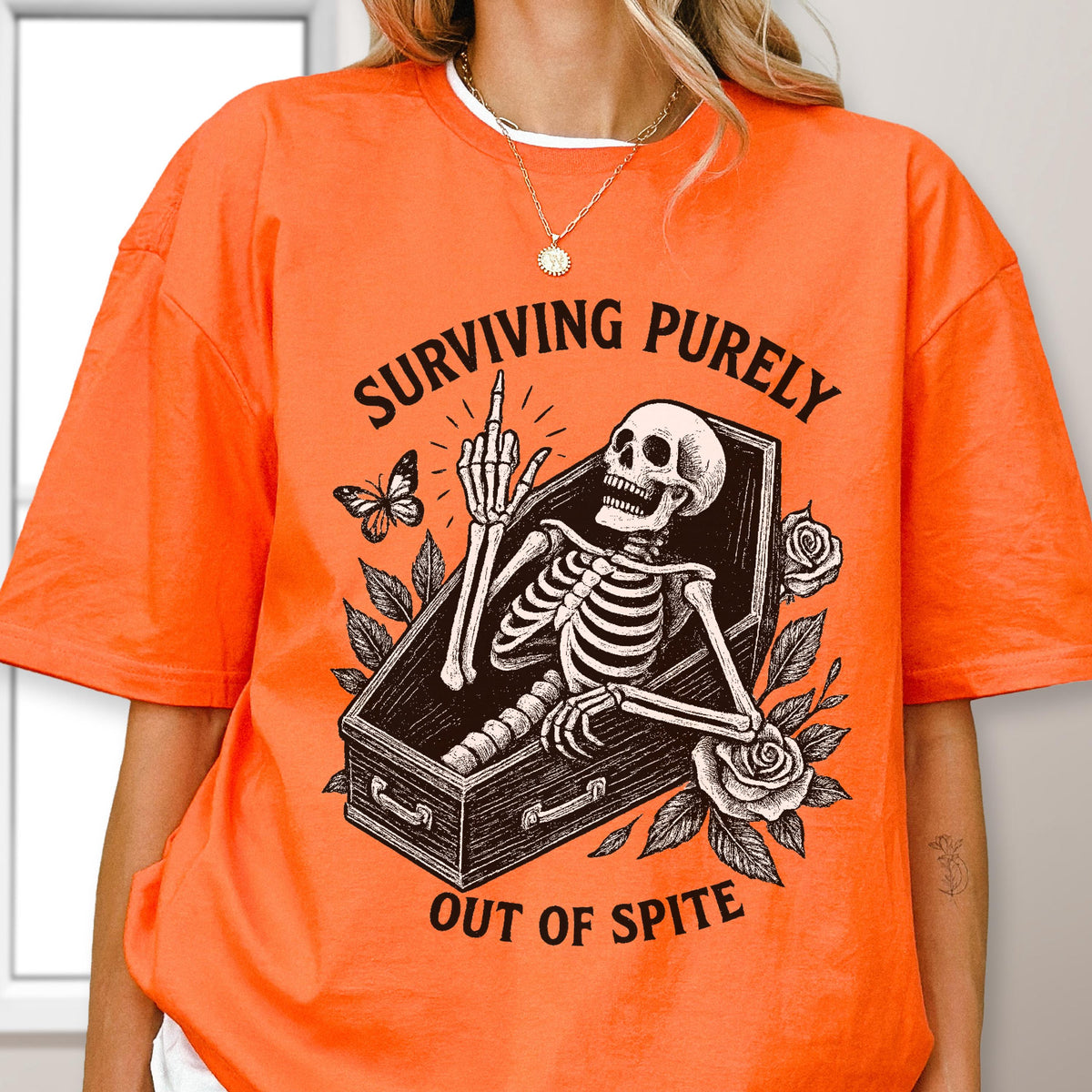 May include: An orange t-shirt featuring a black graphic print of a skeleton inside a coffin, giving the middle finger. The text "SURVIVING PURELY" is arched above the coffin, and "OUT OF SPITE" is printed below, surrounded by roses and a butterfly.