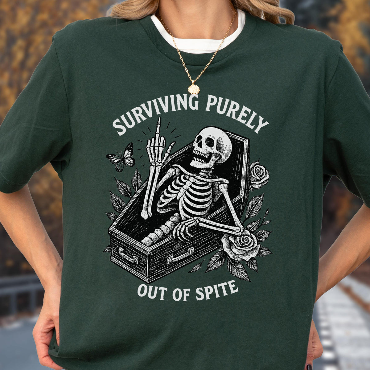 May include: A dark green t-shirt featuring a white graphic of a skeleton inside a coffin, giving the middle finger. The design includes roses, a butterfly, and the text "SURVIVING PURELY OUT OF SPITE" printed in a curved white font above and below.