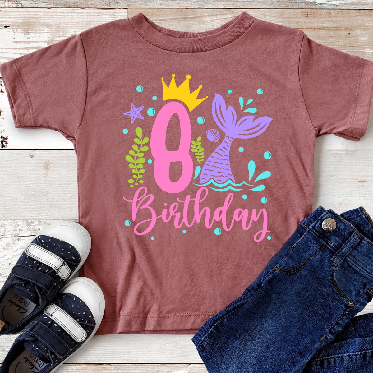 May include: A mauve-colored t-shirt with a colorful mermaid-themed design for an 8th birthday. The design includes a pink number 8, a mermaid tail, a crown, and the word "Birthday" in pink script. Blue jeans and navy blue shoes are next to the shirt.