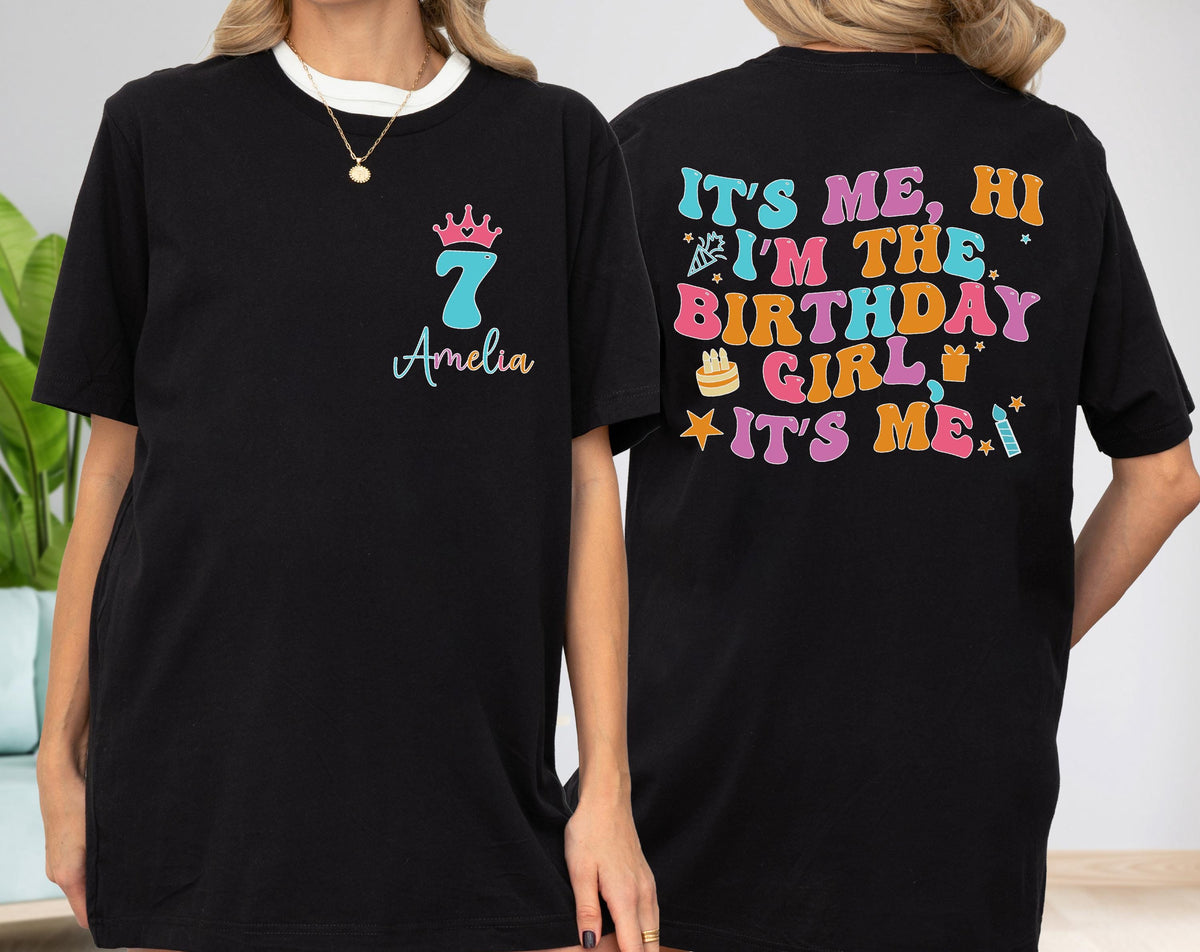 May include: Black t-shirt with a white neckline. The front features a pink crown, the number 7 in teal, and the name "Amelia" in script. The back of the shirt displays the text "IT'S ME, HI I'M THE BIRTHDAY GIRL IT'S ME!" in various colors.