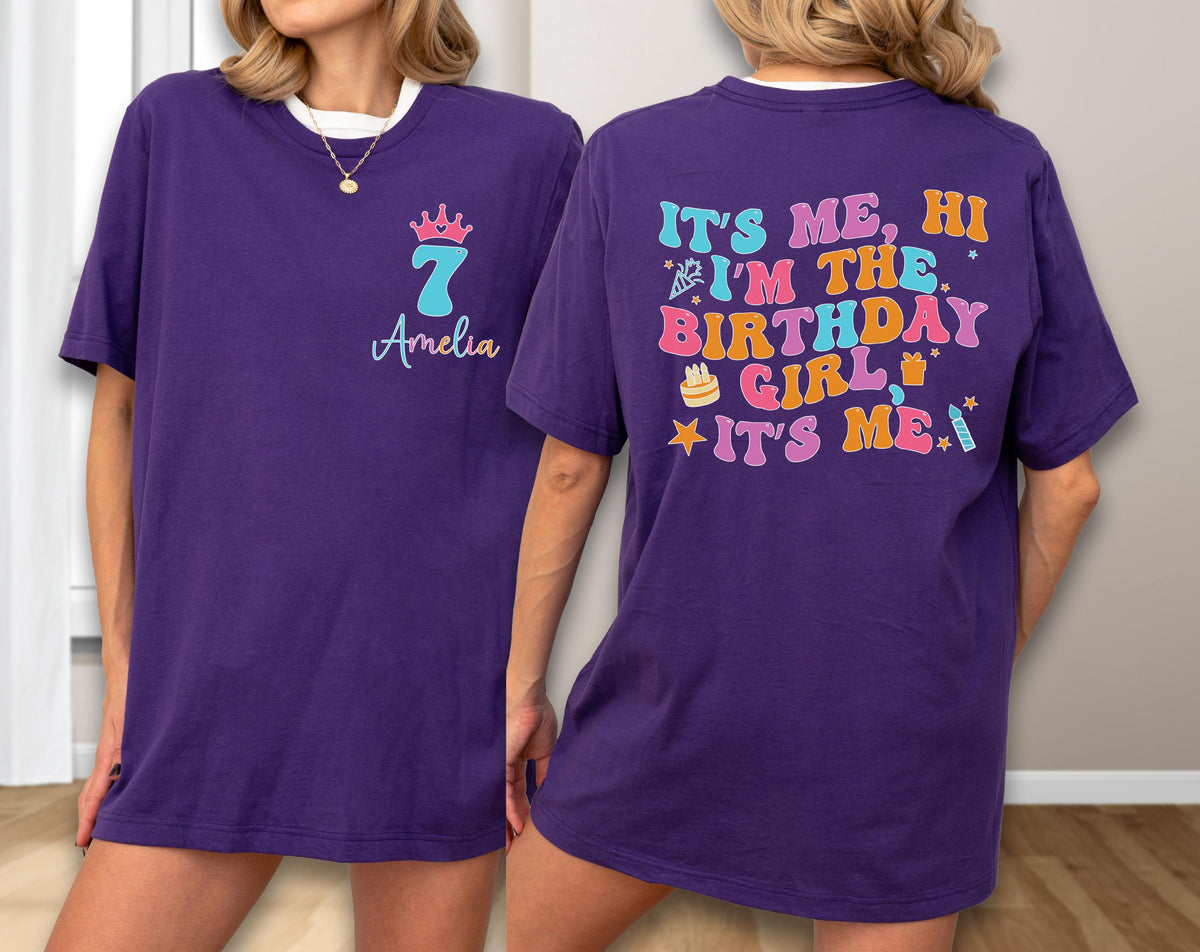 May include: A purple t-shirt featuring a front design with a pink crown, the number 7, and the name Amelia. The back displays the colorful text IT'S ME, HI I'M THE BIRTHDAY GIRL IT'S ME, accompanied by illustrations of a cake, gift, and candle.
