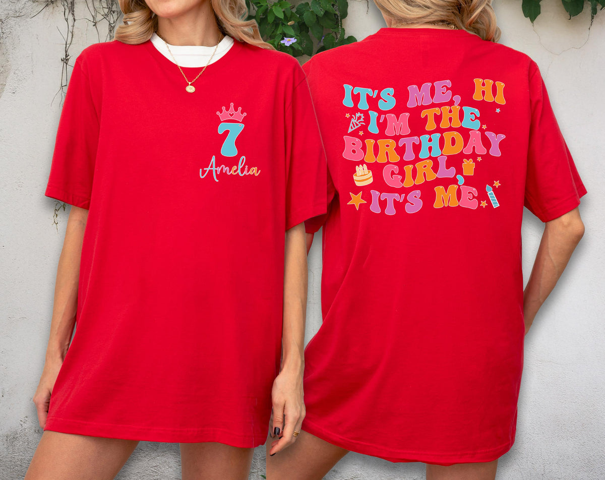May include: A red short-sleeve t-shirt featuring a front graphic with a crown, the number 7, and the name Amelia. The back displays colorful text reading IT'S ME, HI I'M THE BIRTHDAY GIRL IT'S ME, accompanied by small party-themed icons.