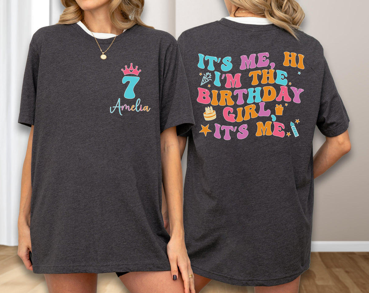 May include: A dark charcoal gray t-shirt featuring a front graphic with a pink crown, the number 7, and the name Amelia. The back displays colorful text reading IT'S ME, HI I'M THE BIRTHDAY GIRL IT'S ME, accompanied by icons of a cake, gift, and candle.
