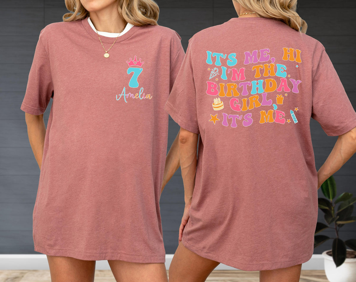 May include: A dusty rose t-shirt featuring a front graphic with a light blue number seven, a pink crown, and the name Amelia. The back displays colorful, retro-style text reading IT'S ME, HI I'M THE BIRTHDAY GIRL IT'S ME, with small birthday icons.