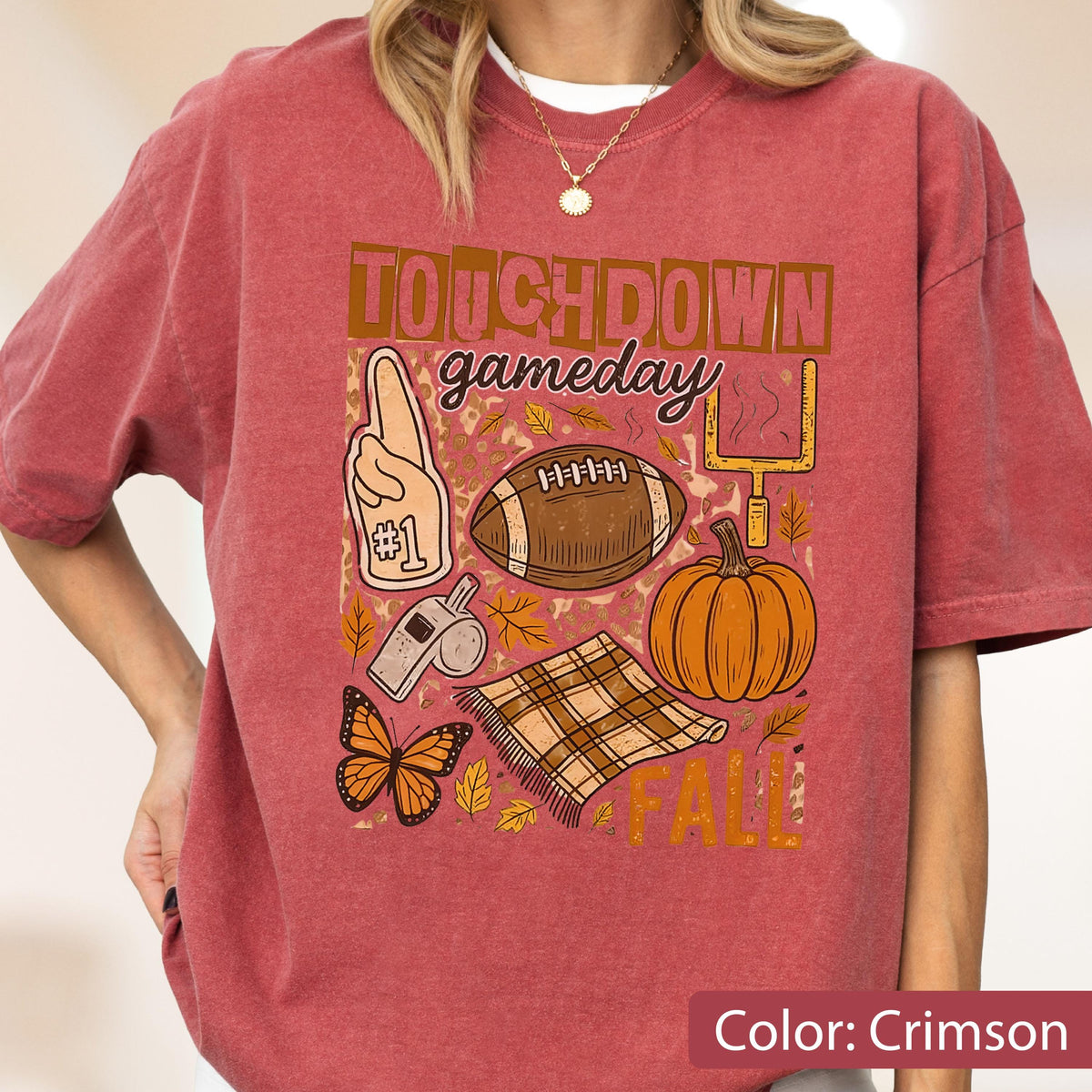 May include: Crimson-colored t-shirt with a football-themed graphic. The design includes a football, pumpkin, butterfly, and the words "TOUCHDOWN gameday FALL". The shirt has short sleeves and a relaxed fit.