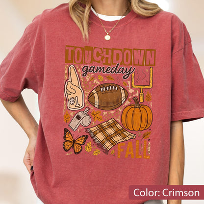May include: Crimson-colored t-shirt with a football-themed graphic. The design includes a football, pumpkin, butterfly, and the words "TOUCHDOWN gameday FALL". The shirt has short sleeves and a relaxed fit.