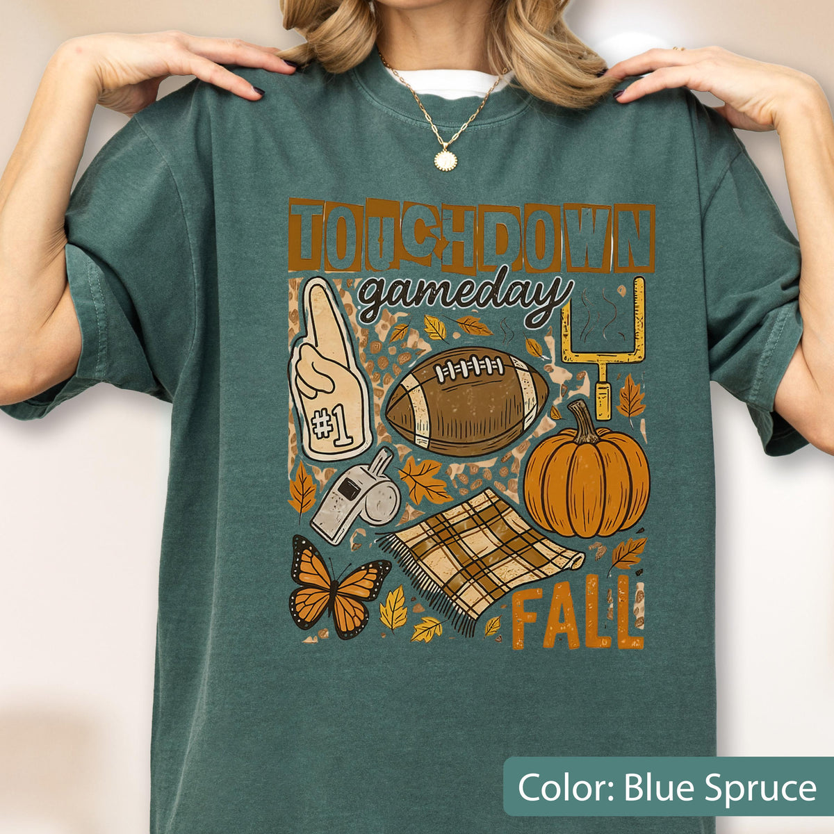 May include: Blue spruce t-shirt with a fall-themed graphic. The design includes a football, pumpkin, and the words "Touchdown Gameday Fall". Other elements include a foam finger, a whistle, a butterfly, and a blanket.