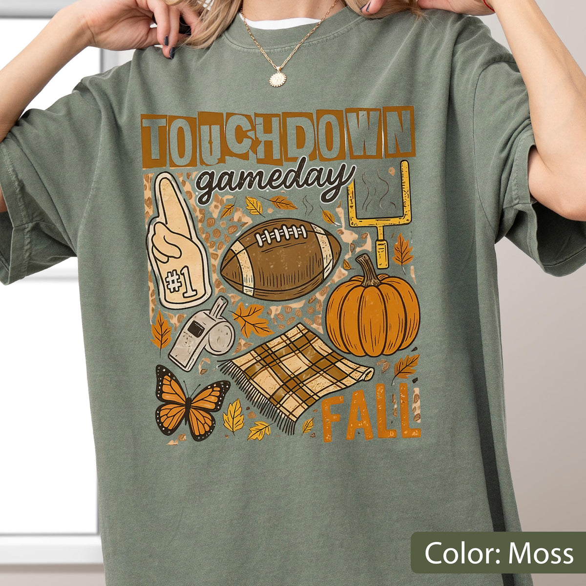 May include: A moss green t-shirt with a football-themed graphic. The design includes a football, pumpkin, foam finger, and the words "TOUCHDOWN gameday FALL". The shirt is a comfortable, casual style.