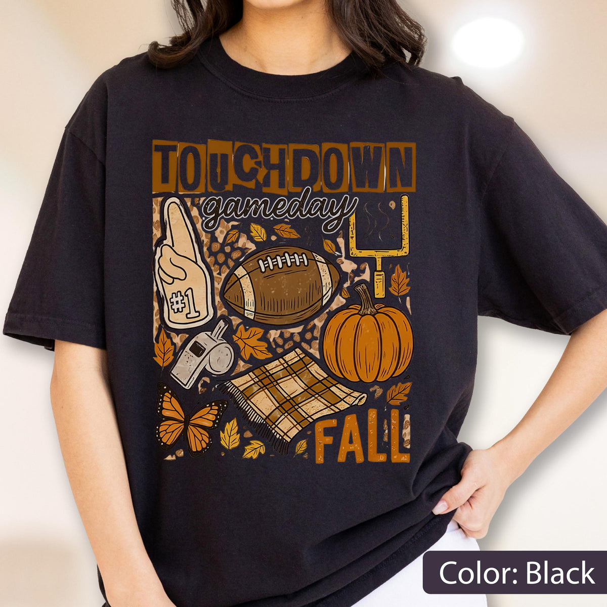 May include: Black t-shirt with a fall-themed graphic. The design includes a football, pumpkin, leaves, and the words "TOUCHDOWN gameday FALL". Additional elements include a foam finger, whistle, and a butterfly.