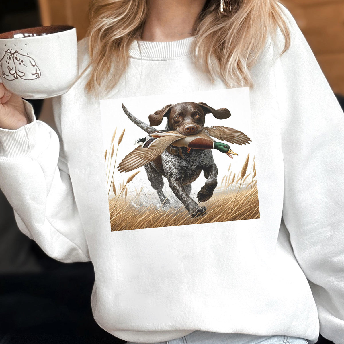 May include: White sweatshirt featuring a graphic of a brown and white dog running through tall grass, carrying a duck in its mouth. The dog is a hunting breed, and the image has a natural, outdoor theme. The sweatshirt is a casual, comfortable style.