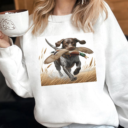 May include: White sweatshirt featuring a graphic of a brown and white dog running through tall grass, carrying a duck in its mouth. The dog is a hunting breed, and the image has a natural, outdoor theme. The sweatshirt is a casual, comfortable style.