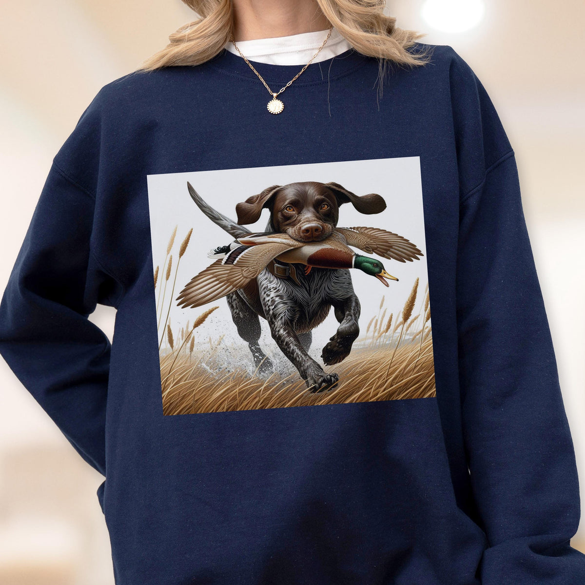 May include: Navy blue sweatshirt featuring a graphic of a hunting dog with a duck in its mouth, running through tall grass. The dog is brown and white, and the duck has brown and green feathers. The background is a light beige.