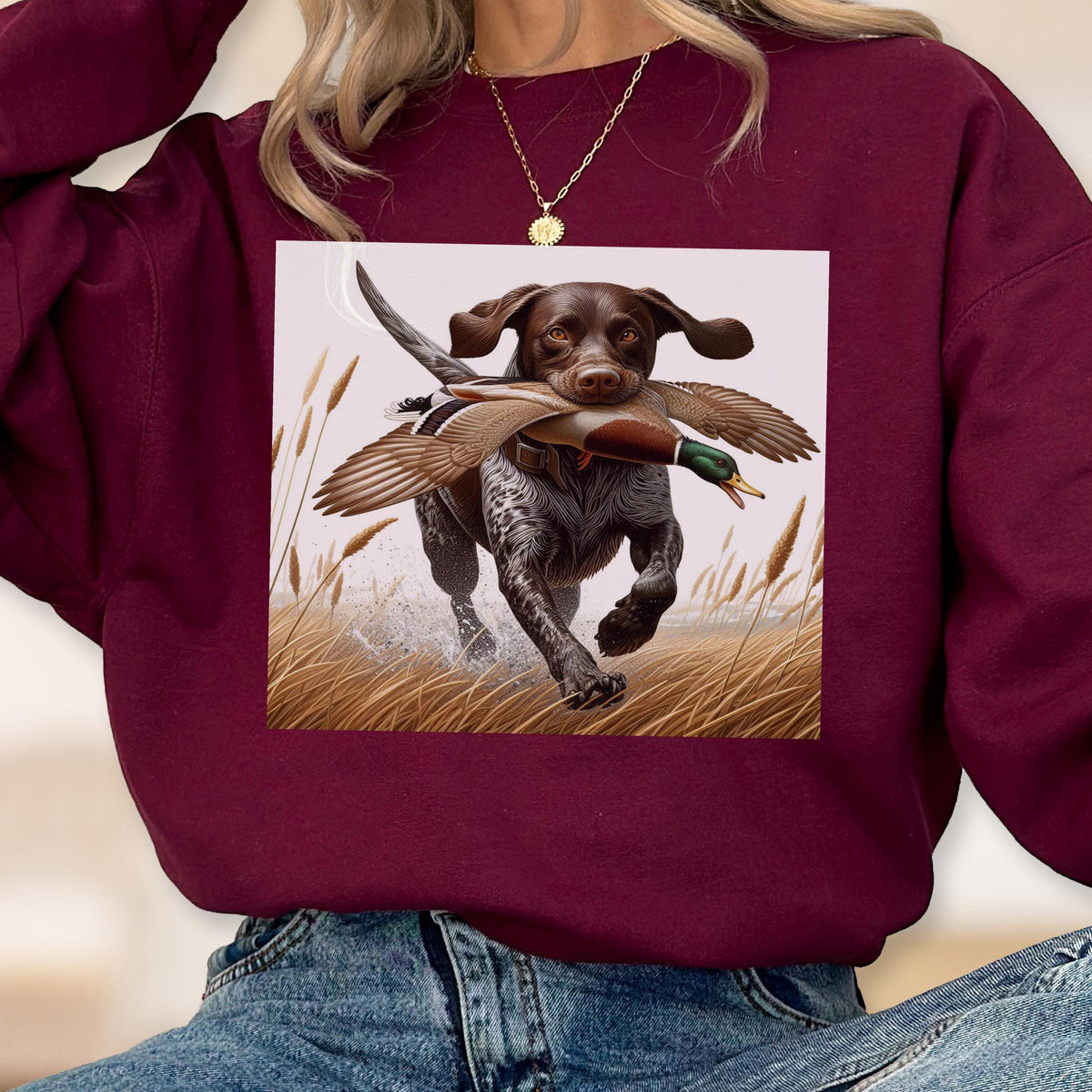 May include: Burgundy sweatshirt featuring a digital illustration of a hunting dog with a duck in its mouth, running through tall grass. The dog is brown with black spots. The duck has brown, green, and white feathers. The background is a light beige.