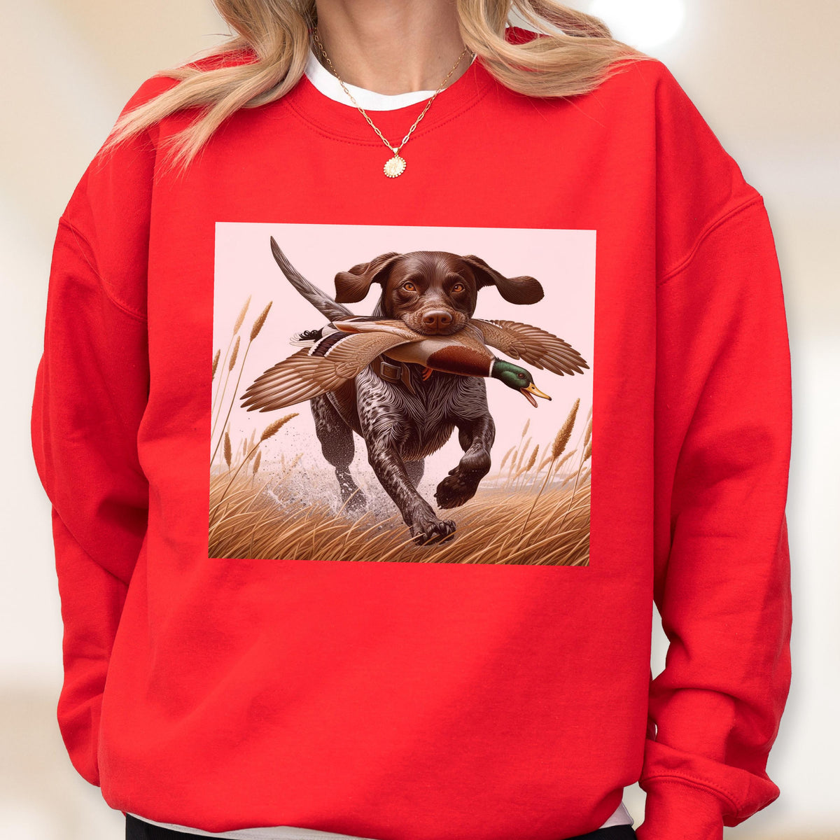 May include: Red sweatshirt featuring a graphic of a brown and white dog running through water with a duck in its mouth. The dog is in a field of tall grass. The sweatshirt is a solid red color.