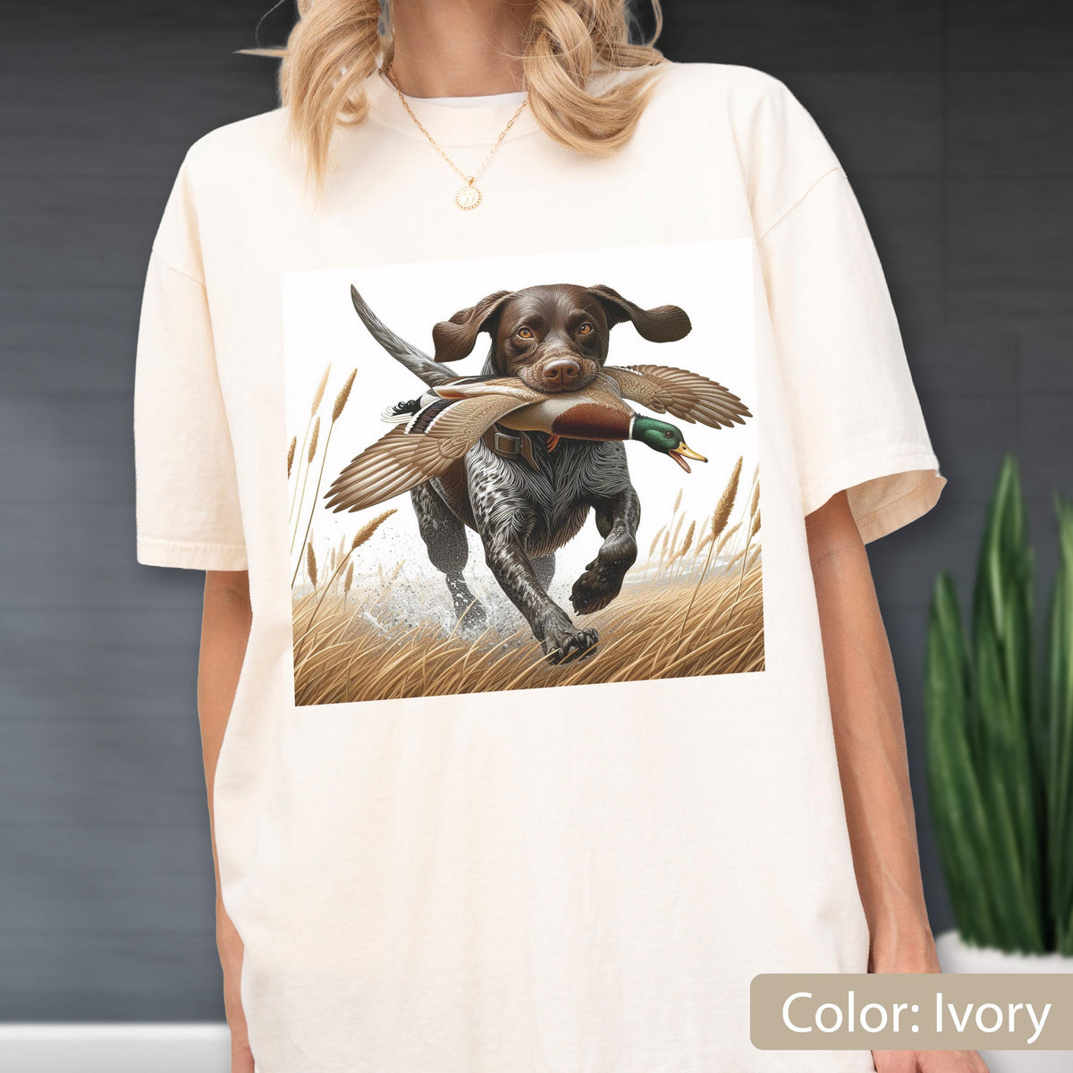 May include: An ivory-colored t-shirt featuring a graphic print of a brown and white hunting dog running through tall grass while carrying a mallard duck in its mouth. The dog has spotted fur and floppy ears, and the scene includes tall reeds.