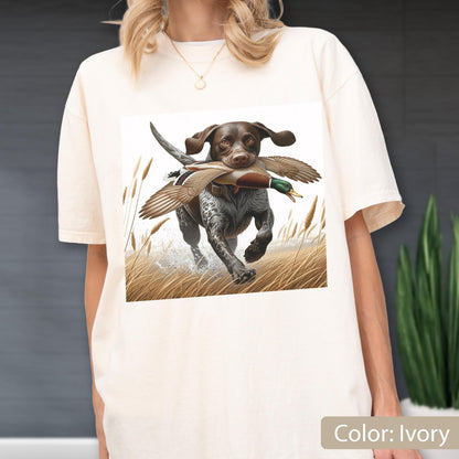 May include: An ivory-colored t-shirt featuring a graphic print of a brown and white hunting dog running through tall grass while carrying a mallard duck in its mouth. The dog has spotted fur and floppy ears, and the scene includes tall reeds.
