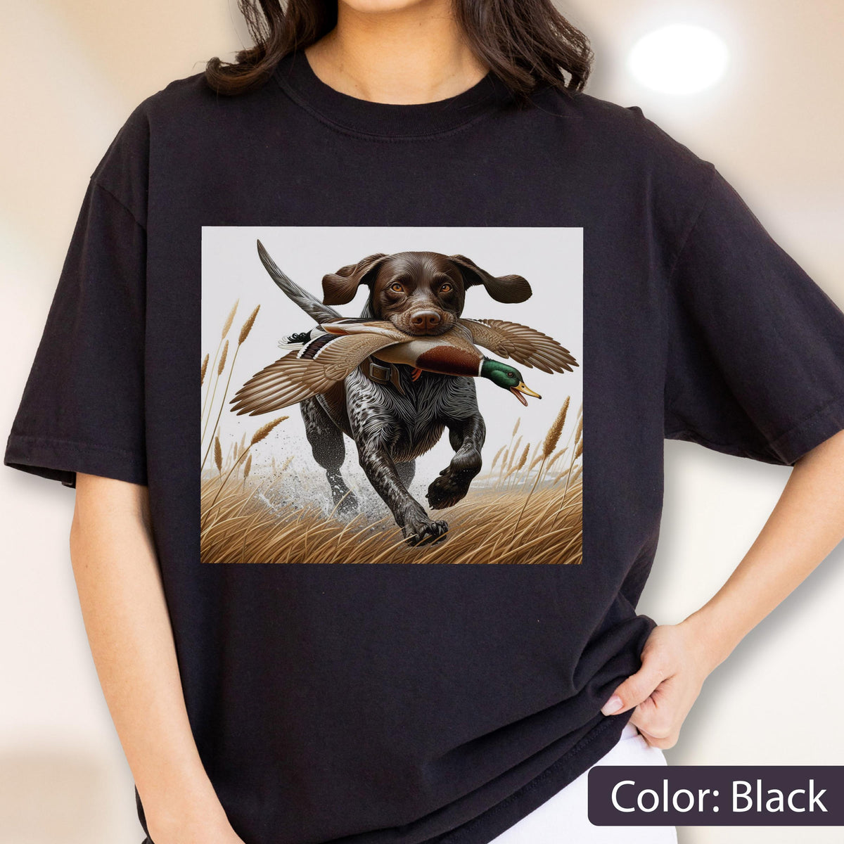 May include: A black short-sleeve t-shirt featuring a graphic print of a brown and white spotted hunting dog running through tall grass while carrying a mallard duck in its mouth. The dog has floppy ears and is depicted in a field setting.