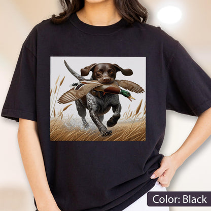 May include: A black short-sleeve t-shirt featuring a graphic print of a brown and white spotted hunting dog running through tall grass while carrying a mallard duck in its mouth. The dog has floppy ears and is depicted in a field setting.