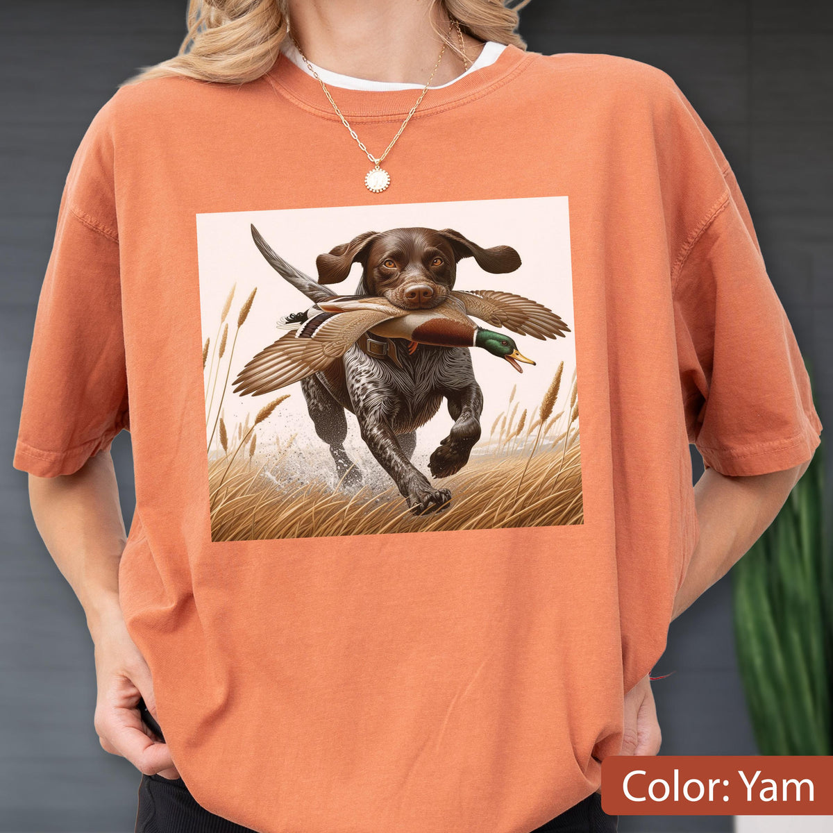 May include: A burnt orange crewneck t-shirt featuring a graphic print of a brown and white spotted hunting dog running through a field of tall grass while carrying a mallard duck in its mouth. The shirt has short sleeves and a relaxed fit.