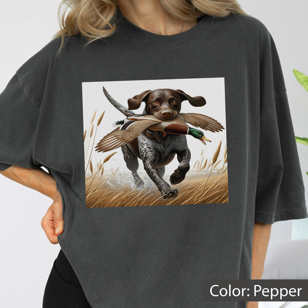May include: A charcoal gray t-shirt featuring a graphic print of a brown and white hunting dog running through a field of tall grass while carrying a mallard duck in its mouth. The dog has spotted fur and the duck displays brown and green feathers.