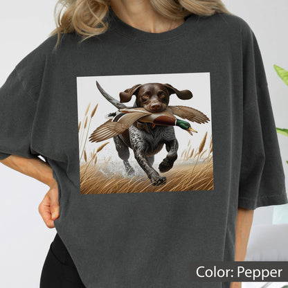 May include: A charcoal gray t-shirt featuring a graphic print of a brown and white hunting dog running through a field of tall grass while carrying a mallard duck in its mouth. The dog has spotted fur and the duck displays brown and green feathers.