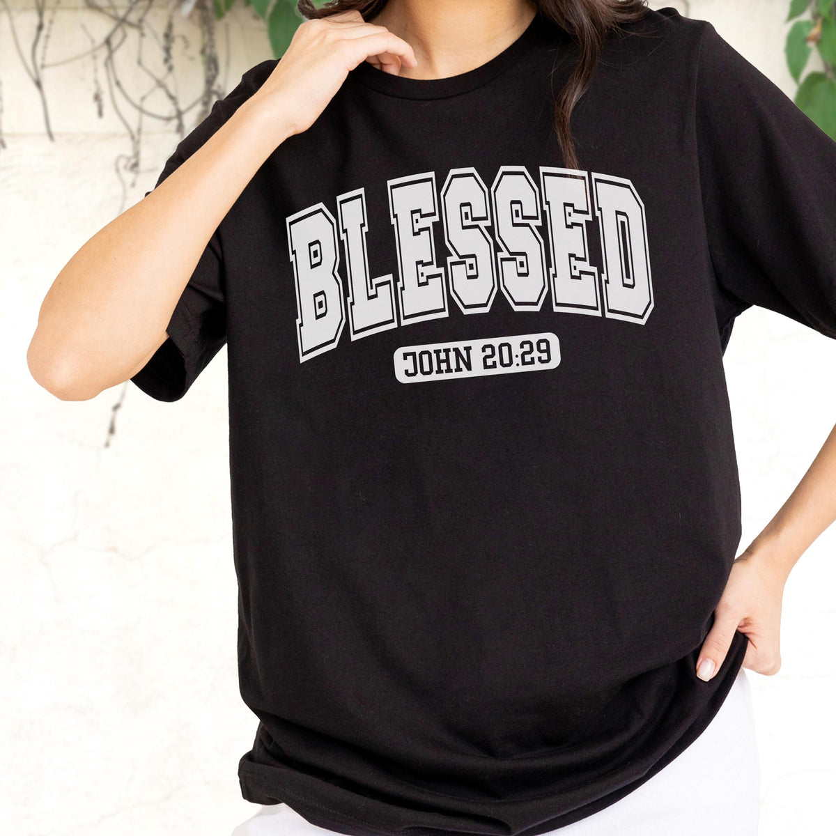 May include: A black short-sleeved t-shirt featuring the word BLESSED in a large, grey, collegiate-style font across the chest. Below the main text, the phrase JOHN 20:29 is printed in a smaller, grey, rectangular block with rounded corners.
