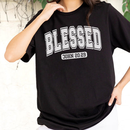 May include: A black short-sleeved t-shirt featuring the word BLESSED in a large, grey, collegiate-style font across the chest. Below the main text, the phrase JOHN 20:29 is printed in a smaller, grey, rectangular block with rounded corners.