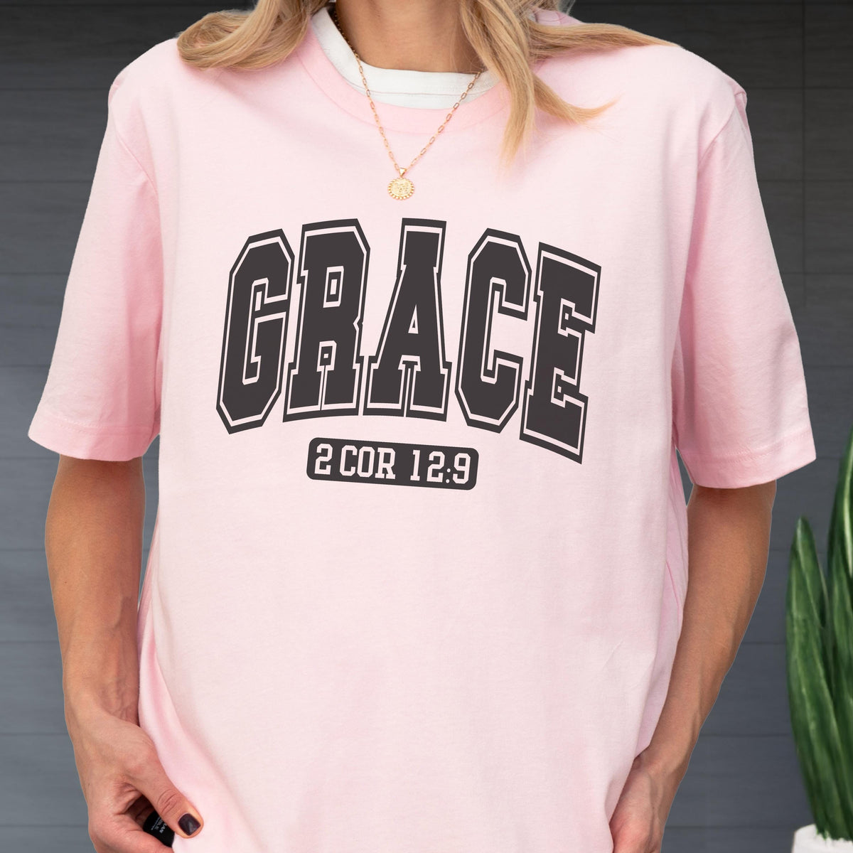 May include: A light pink short-sleeved t-shirt featuring the word GRACE in large black collegiate-style lettering across the chest. Below the word, the text 2 COR 12:9 is printed in a smaller black rectangular box with rounded corners.