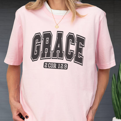 May include: A light pink short-sleeved t-shirt featuring the word GRACE in large black collegiate-style lettering across the chest. Below the word, the text 2 COR 12:9 is printed in a smaller black rectangular box with rounded corners.