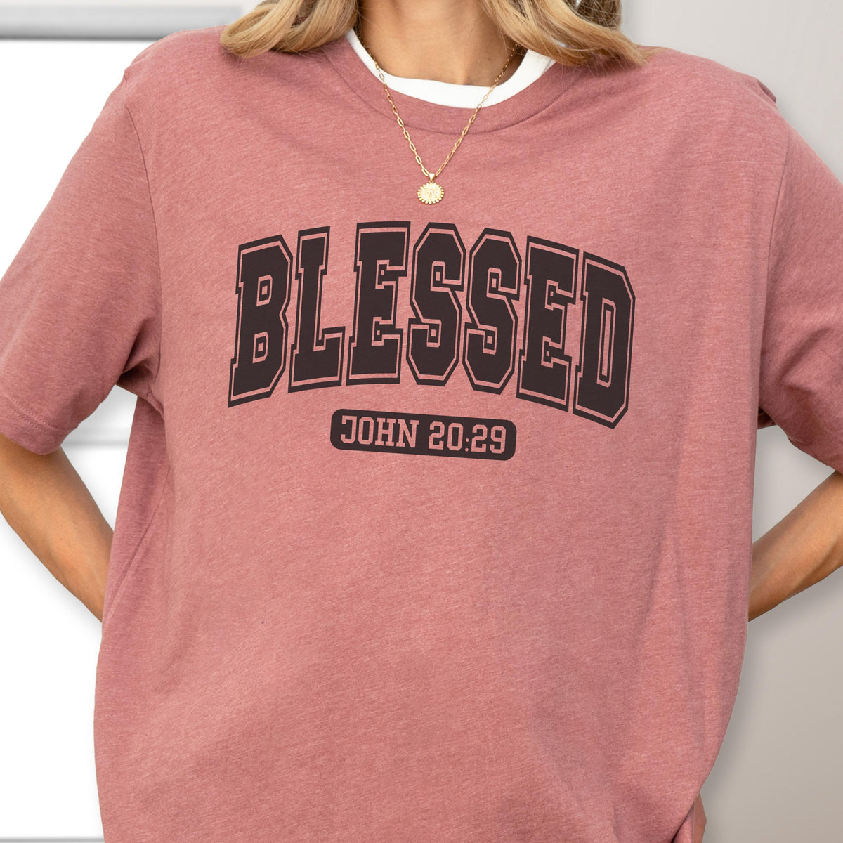 May include: A dusty rose short-sleeve t-shirt featuring the word BLESSED in a large black collegiate-style font across the chest. Below the word, the text JOHN 20:29 is printed in a smaller black rectangular box. The shirt has a crew neckline.