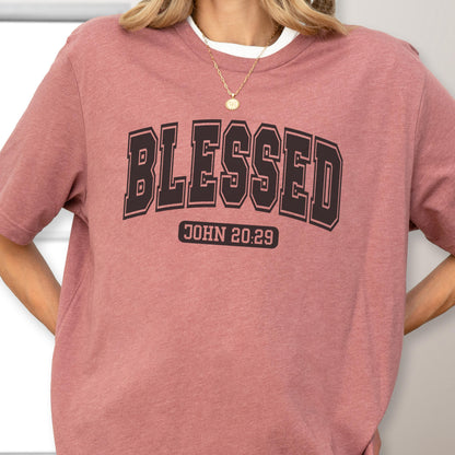 May include: A dusty rose short-sleeve t-shirt featuring the word BLESSED in a large black collegiate-style font across the chest. Below the word, the text JOHN 20:29 is printed in a smaller black rectangular box. The shirt has a crew neckline.