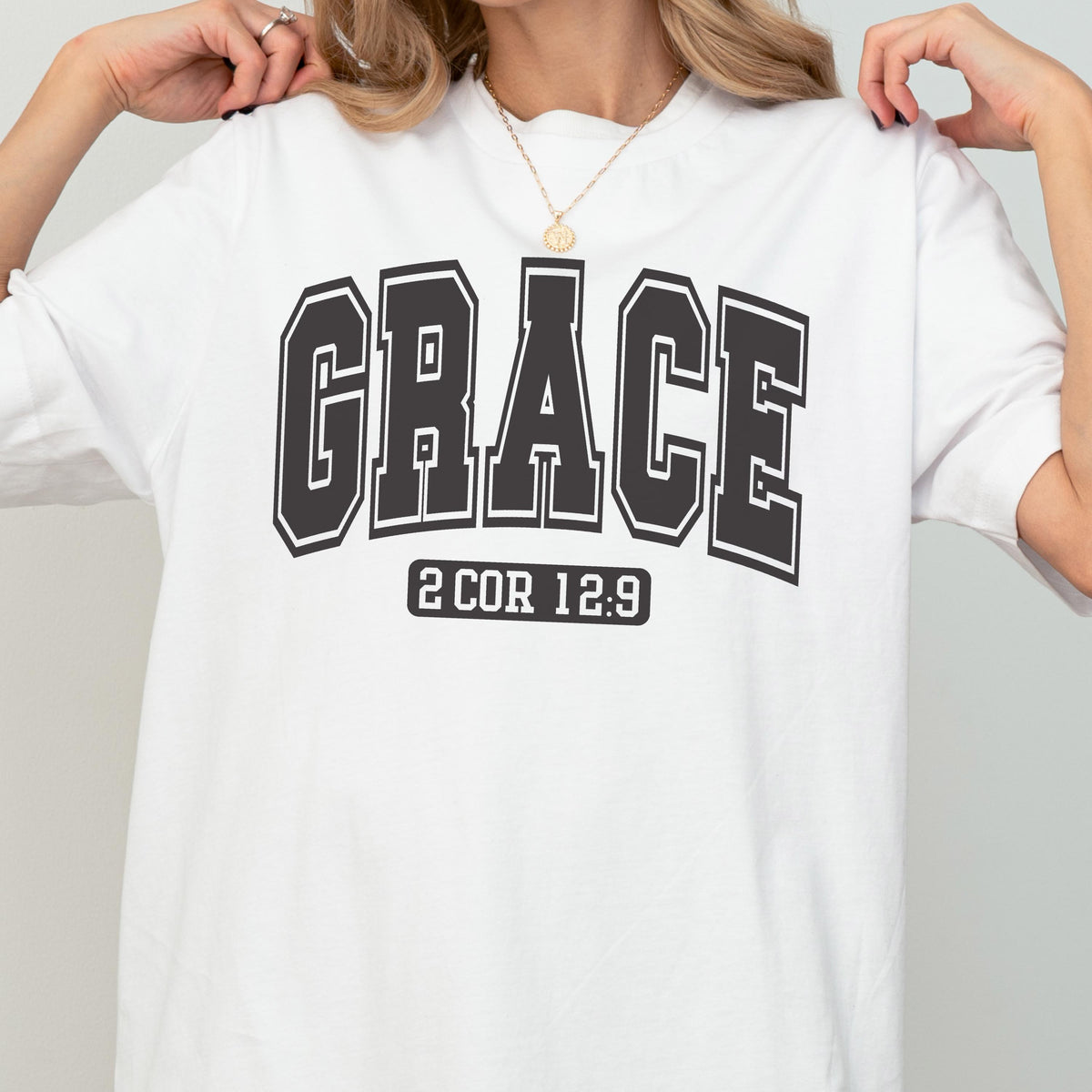 May include: A white short-sleeved t-shirt featuring the word GRACE in large, bold, black collegiate-style lettering across the chest. Below the word, the text 2 COR 12:9 is printed in a smaller, rectangular black block with white lettering.