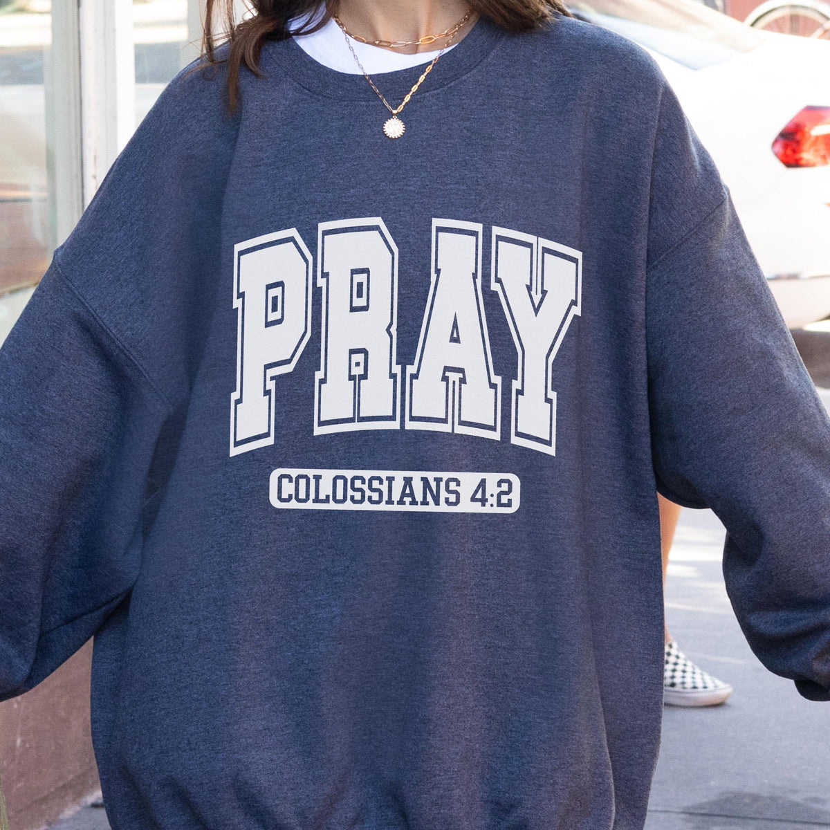 May include: A dark heather navy blue crewneck sweatshirt featuring the word PRAY in large white block letters with a white outline, followed by the text COLOSSIANS 4:2 in a smaller white font centered below the main word on the chest area.
