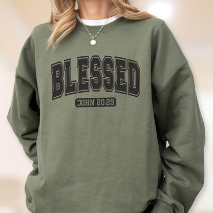 May include: An olive green crewneck sweatshirt featuring the word BLESSED in a large black collegiate-style font arched across the chest, with the text JOHN 20:29 printed in a smaller black rectangular block directly underneath the main word.