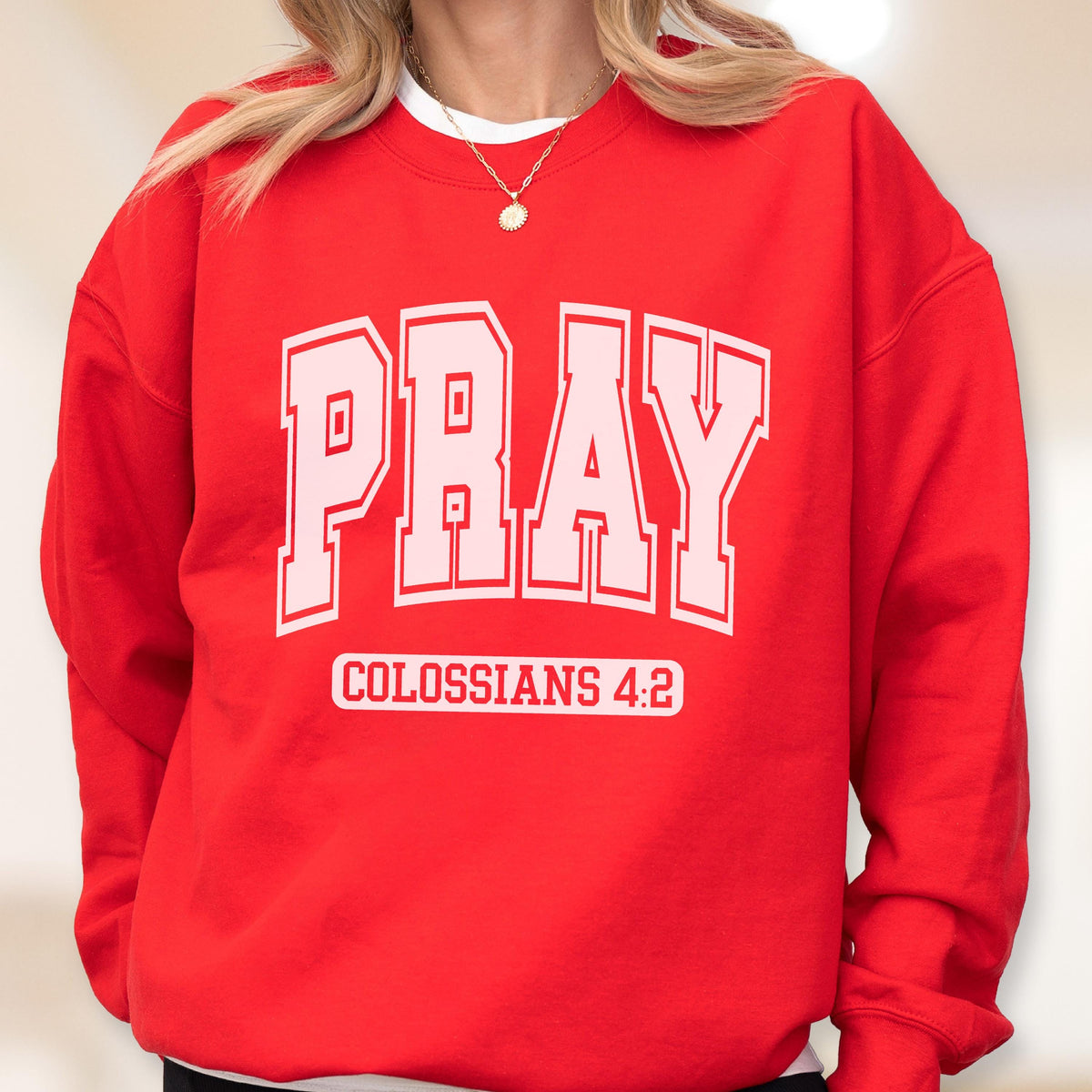 May include: A bright red crewneck sweatshirt featuring the word PRAY in large, light pink block letters with a white outline. Below the word, the text COLOSSIANS 4:2 is printed in white capital letters inside a rectangular white border.