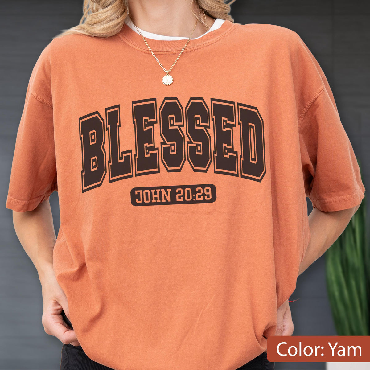 May include: A yam-colored short-sleeve t-shirt featuring the word BLESSED in large black collegiate-style lettering across the chest, with the text JOHN 20:29 printed in a smaller black rectangular box directly underneath the main word.
