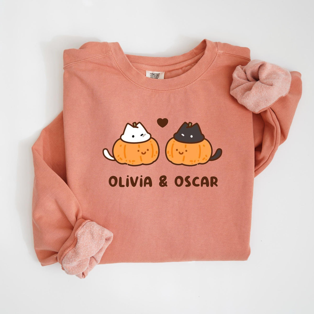 May include: A coral-colored sweatshirt featuring a graphic of two cartoon cats inside pumpkins, with a heart above them. The text "OLIVIA & OSCAR" is printed below the pumpkins. A matching scrunchie is on the right side.
