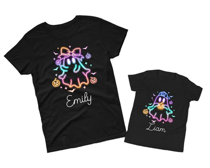 May include: Two black t-shirts with Halloween-themed designs. One shirt features a ghost with a bow, pumpkins, and the name "Emily." The other shirt has a ghost wearing a cap, pumpkins, and the name "Liam." The designs use neon colors.