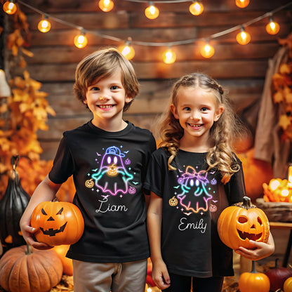 May include: Two children wearing black Halloween t-shirts with neon ghost designs and holding carved pumpkins. The shirts have the names "Liam" and "Emily" printed on them. The background includes autumn foliage and string lights.