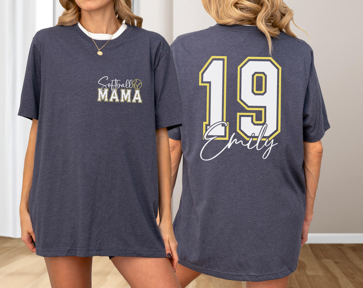 May include: Dark gray t-shirt with white trim. The front features "Softball MAMA" in script and block letters with a softball graphic. The back displays the number "19" in white with a gold outline, and the name "Emily" in script.