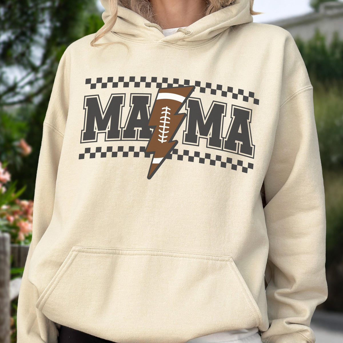 May include: A beige hoodie with a front graphic that says "MAMA" in dark gray block letters, with a brown football-shaped lightning bolt in the center. The design is framed by a black and white checkered pattern.