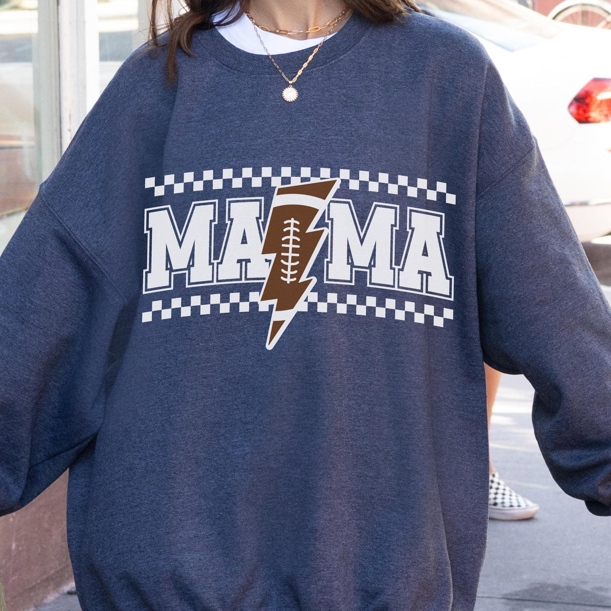 May include: Dark blue sweatshirt with the word "MAMA" in white block letters, a brown football-shaped lightning bolt graphic, and a white and black checkered border. The sweatshirt is a casual style.