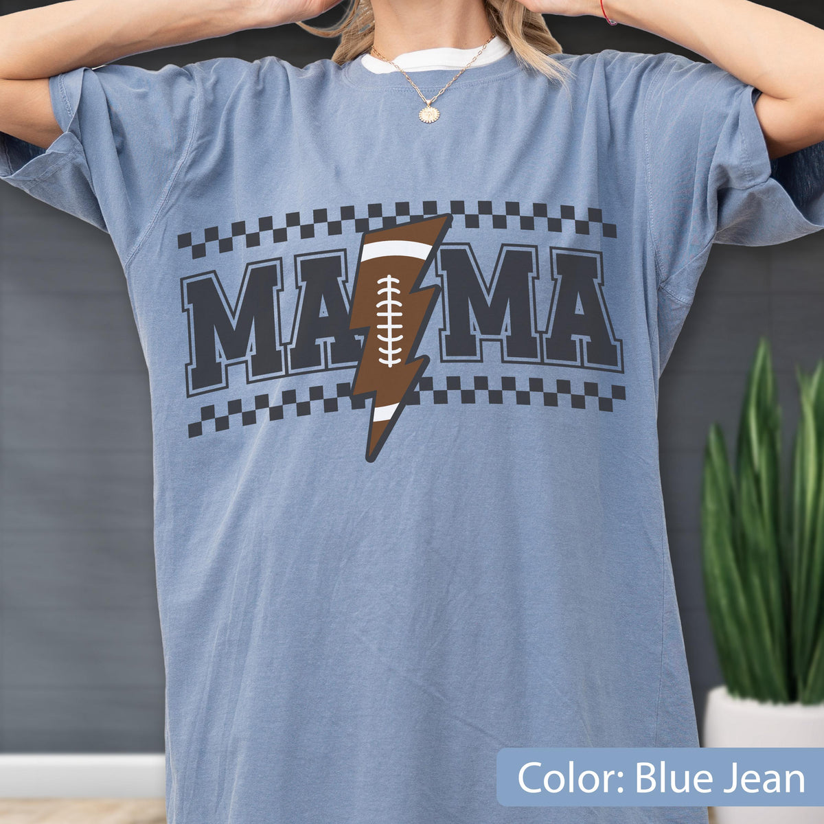 May include: A blue jean colored t-shirt featuring the text MAMA in black block letters. The letter A is replaced by a brown football shaped like a lightning bolt with white laces. The design is framed by black and white checkered borders.