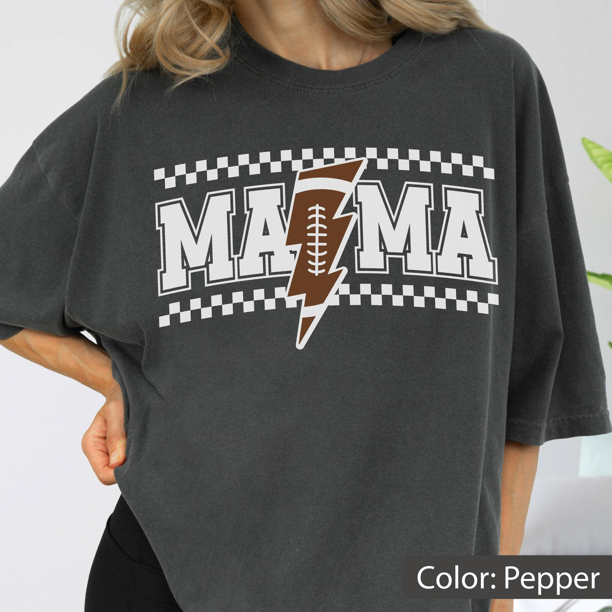 May include: A pepper-colored short-sleeve t-shirt featuring the white text MAMA with a brown and white football-shaped lightning bolt graphic in the center. The design is framed by white and pepper-colored checkered borders above and below the text.