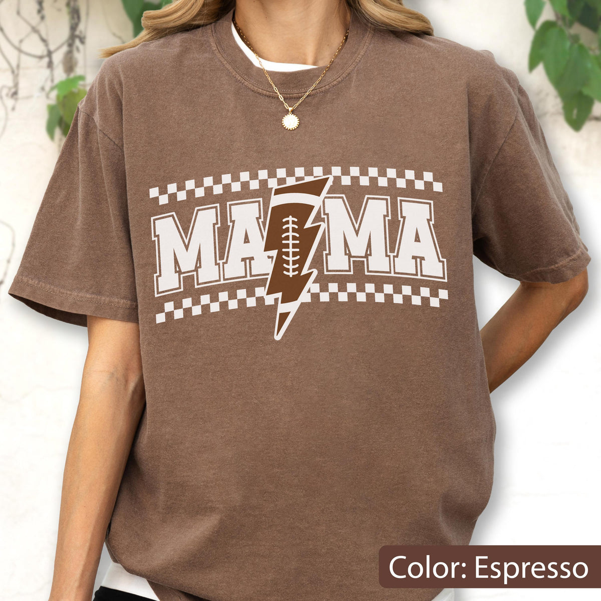 May include: An espresso brown short-sleeve t-shirt featuring a white graphic print that reads MAMA with a brown and white football-shaped lightning bolt replacing the letter A. The design is framed by a white checkered border above and below.
