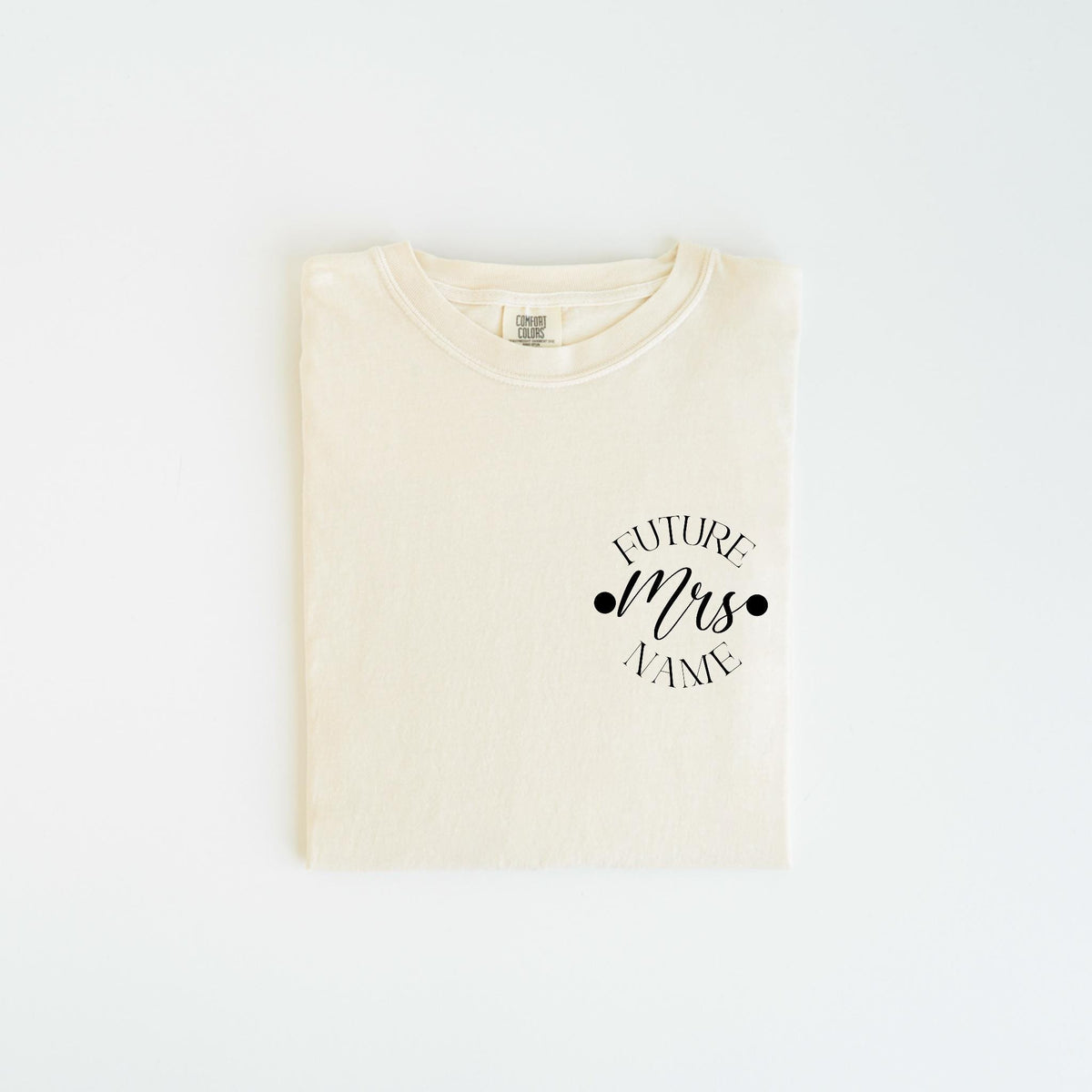 May include: Cream-colored t-shirt with the text "FUTURE Mrs. NAME" in a black script font. The text is centered on the chest, with two black dots on either side of "Mrs.". The shirt is folded neatly on a white surface.