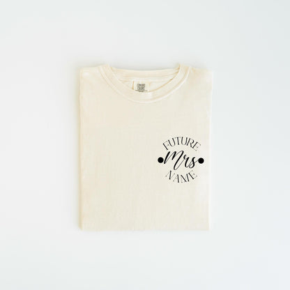 May include: Cream-colored t-shirt with the text "FUTURE Mrs. NAME" in a black script font. The text is centered on the chest, with two black dots on either side of "Mrs.". The shirt is folded neatly on a white surface.