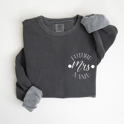 May include: Dark gray sweatshirt with the text "FUTURE Mrs NAME" in white script. The sweatshirt has a crew neck and long sleeves with gray fleece cuffs. The garment is a comfortable, casual style.