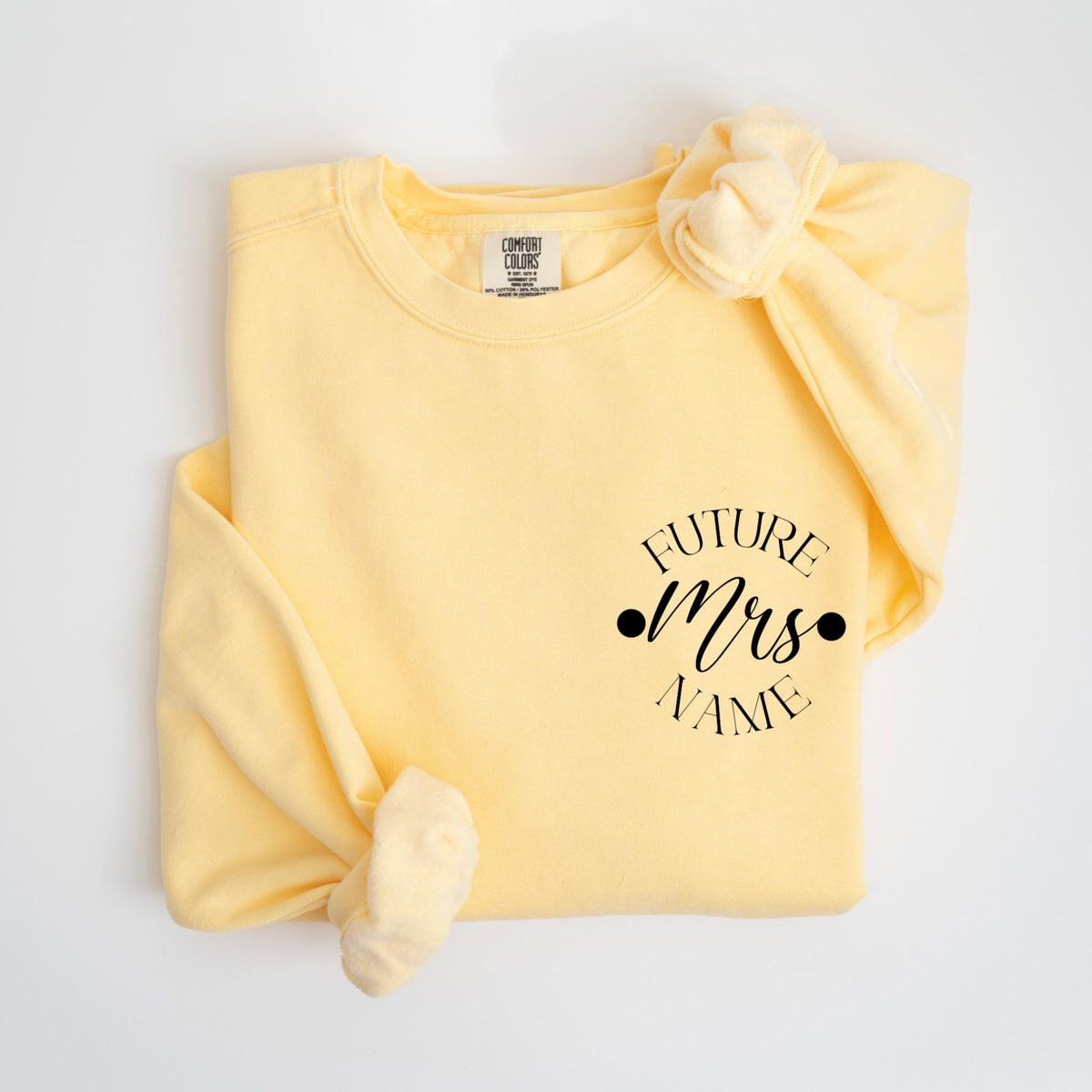 May include: A light yellow sweatshirt with the text "FUTURE Mrs NAME" in black script. The sweatshirt is folded with the sleeves tied in a knot. The Comfort Colors brand tag is visible.